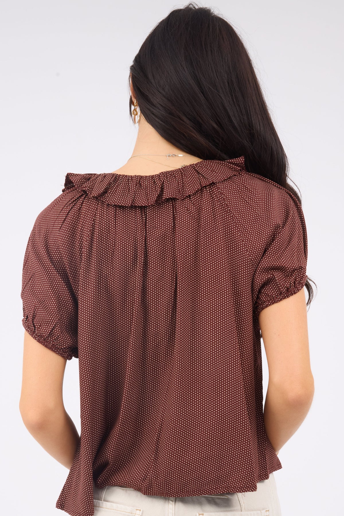 A back view of a woman wearing a loose fitting brown polka dot blouse with a ruffle collar and short puff sleeves.