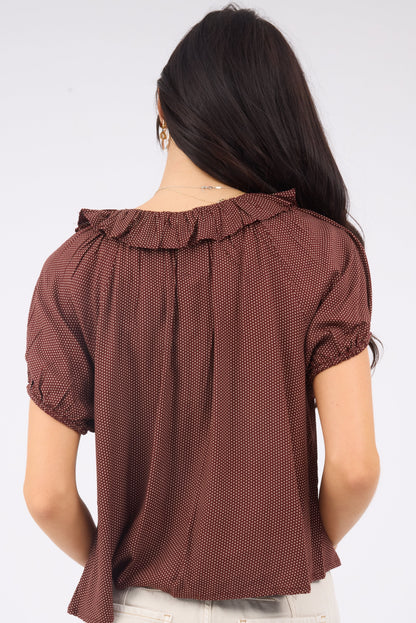 A back view of a woman wearing a loose fitting brown polka dot blouse with a ruffle collar and short puff sleeves.