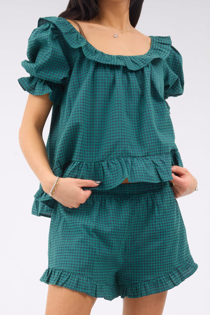 Close up view of a woman weairng a green & navy gingham shorts set with a wide, ruffled scoop neckline, large puff sleeves, ruffle hem.  The shorts have an elastic waistband and ruffled hems.