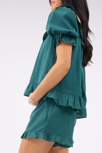 A close up view of a young woman wearing a  green & navy gingham 2 piece shorts & top set ith ruffle details and short sleeves.