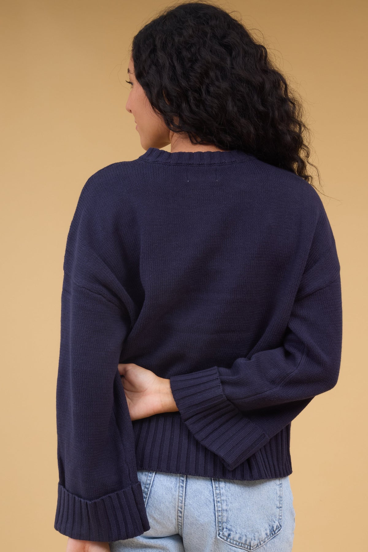 Back view of a woman wearing a loose fitting navy sweater with long sleeves with wide ribbed cuffs.