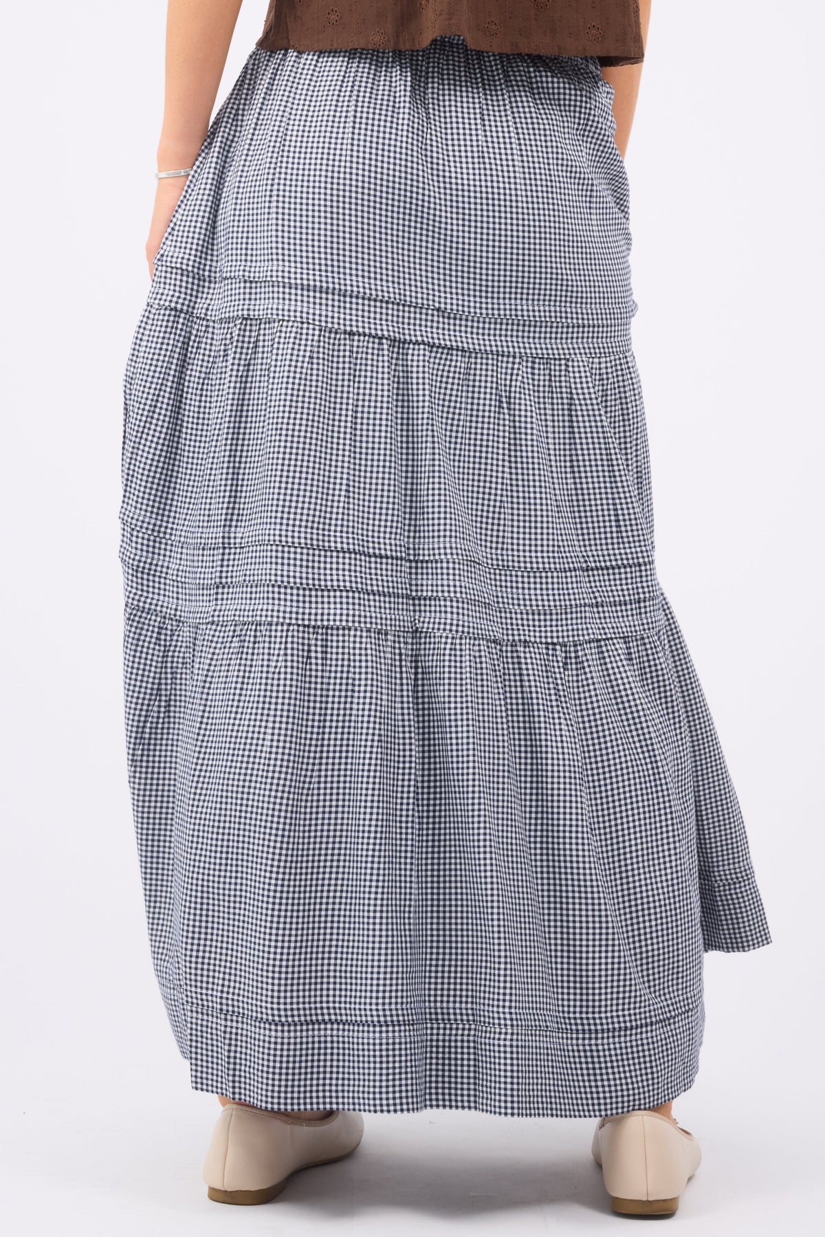 A back view of a woman wearinga navy & white gingham maxi skirt with folded pleats and gathered tiers.