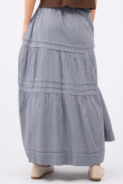 A back view of a woman wearinga navy & white gingham maxi skirt with folded pleats and gathered tiers.
