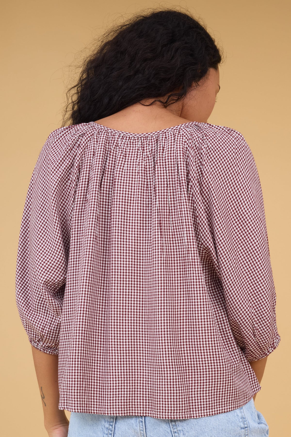 Back view of a woman wearing a burgundy gingham blouse with half length sleeves.