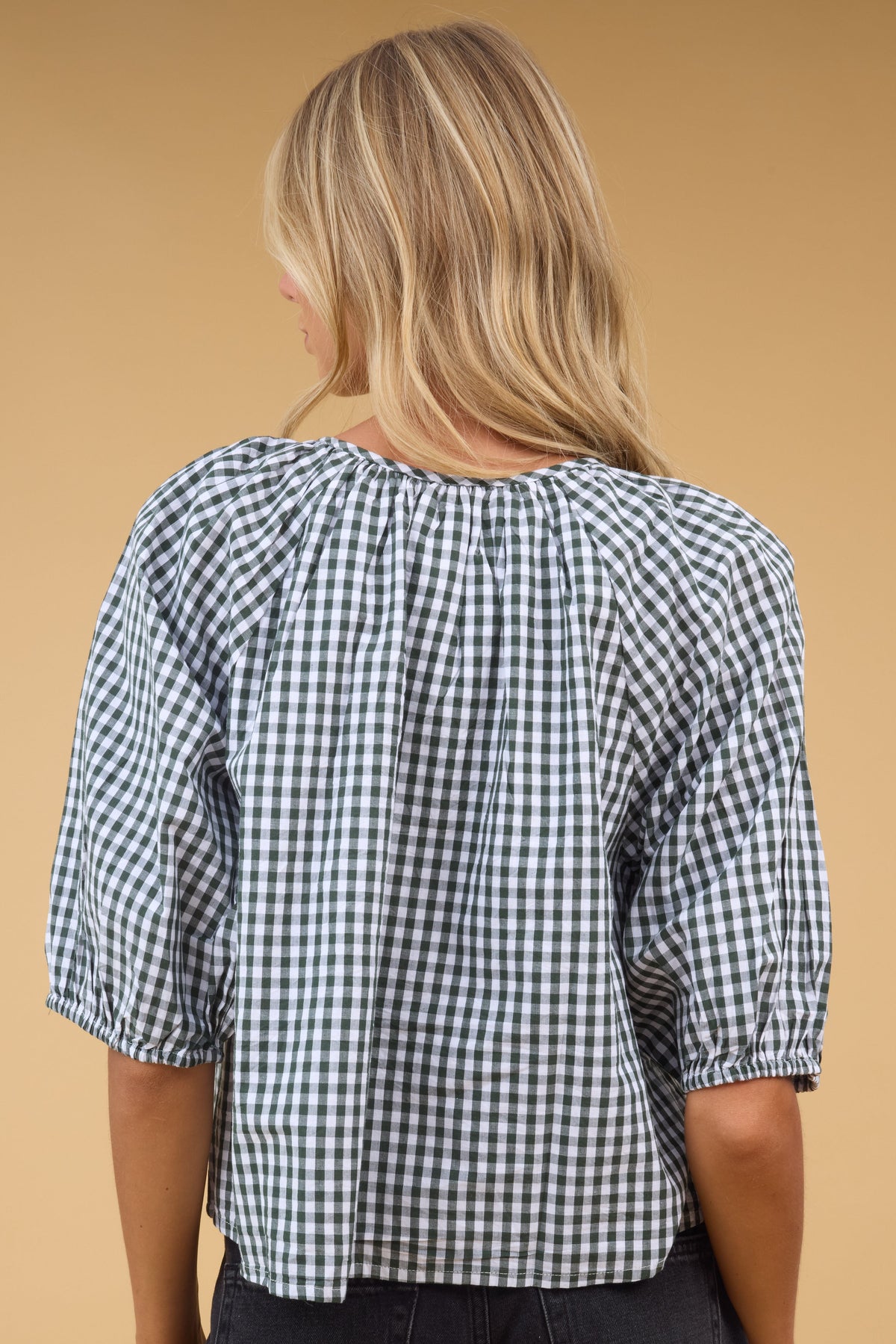 Back view of a woman wearing a loose fitting green gingham blouse with half length sleeves.
