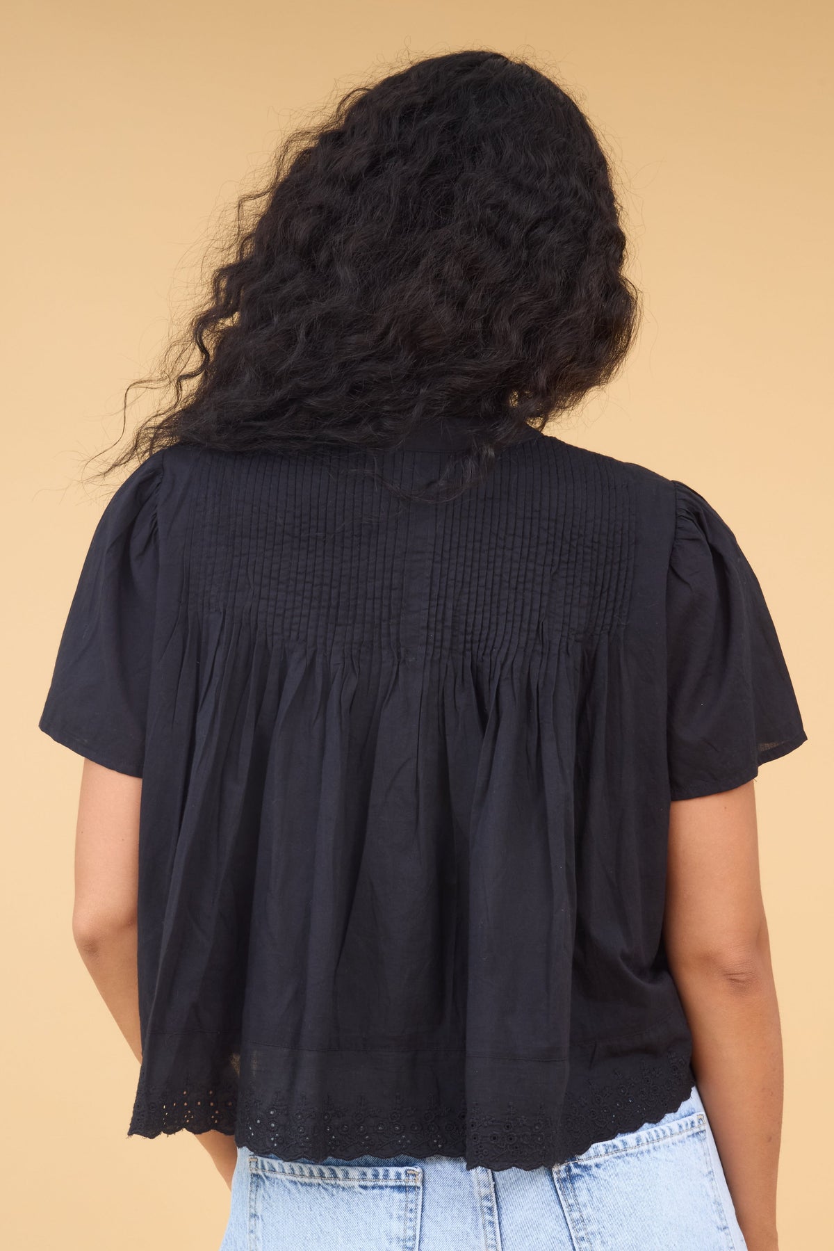Back view of a woman wearing a loose fitting black blouse with short sleeves, pintucks at the shoulders and eyelet trim at the hem.