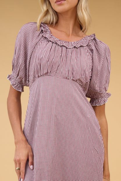 Close view of a woman wearing a brown gingham dress with an empire waist, gathered waist, round ruffled neckline and short puff sleeves.