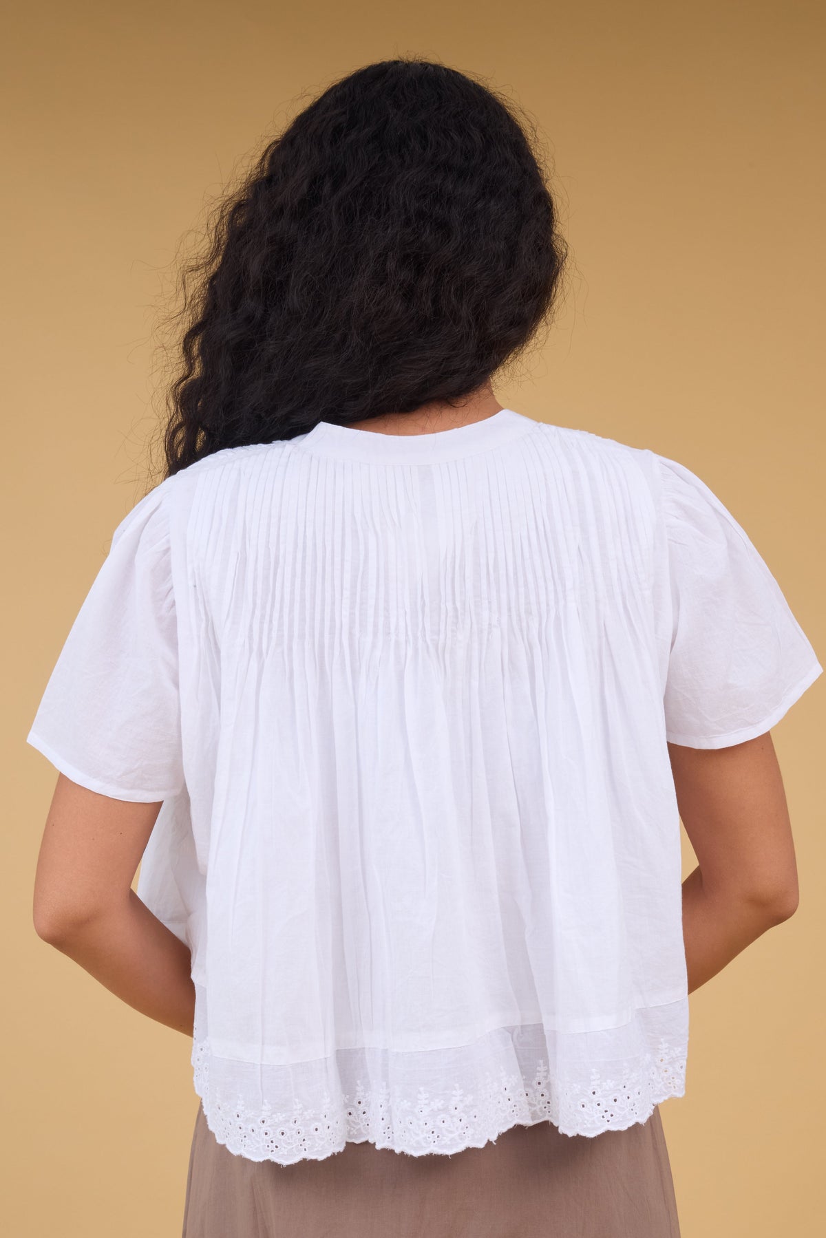 Back view of a woman wearing a white loose fitting blouse with shrot sleeves, pintucks at the shoulders and bottom eyelet lace trim.