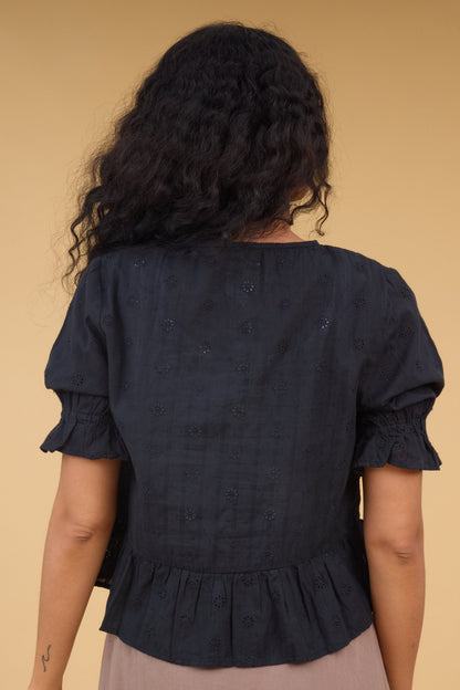 Back view of a young woman wearing a black eyelet, loose fitting blouse with a bottom ruffle and puff sleeves.