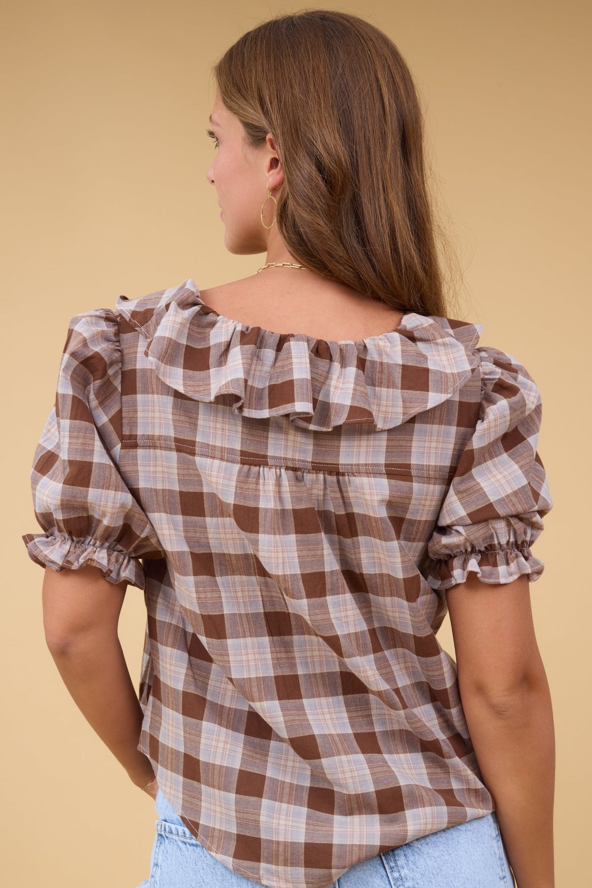 Back view of a woman wearing a loose fitting, brown plaid blouse with a ruffle collar and short puff sleeves.