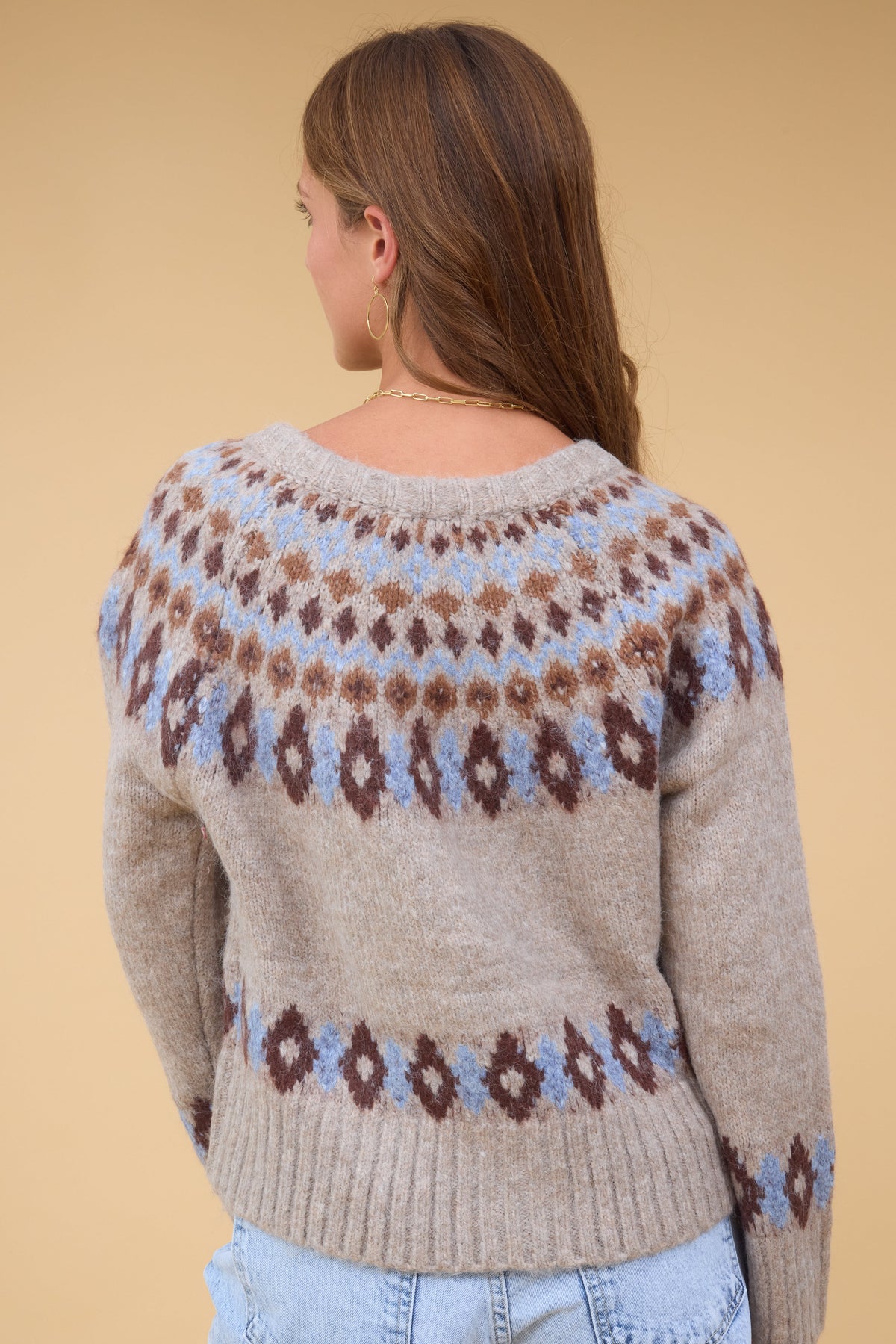 Back view of a young woman wearing a beige long sleeve sweater with a blue & brown nordic pattern.