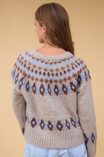 Back view of a young woman wearing a beige long sleeve sweater with a blue & brown nordic pattern.