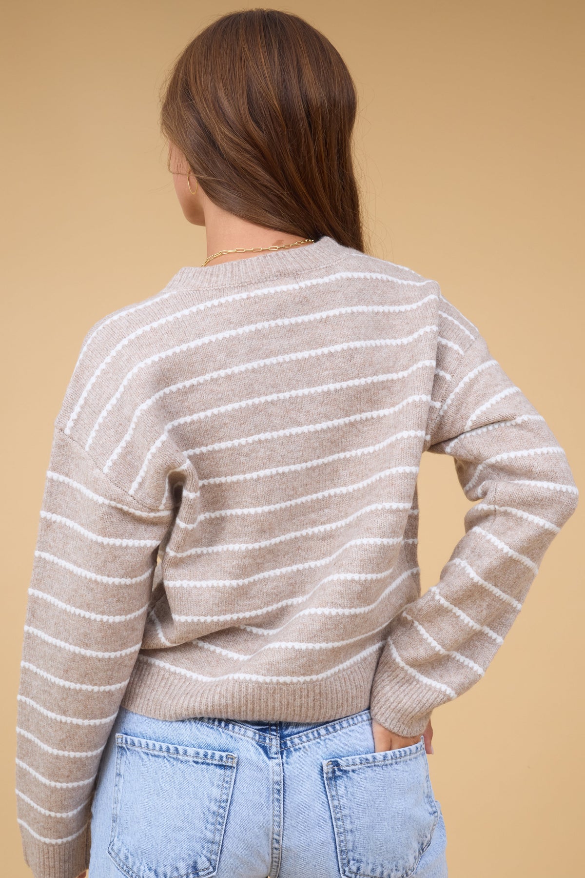 A back view of a young woman wearing a taupe long sleeve cardigan with thin lace textured stripes.