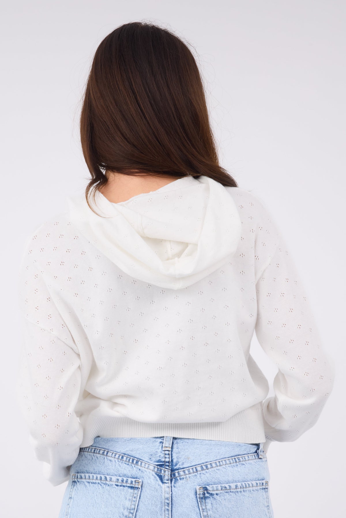 A back view of a young woman wearing a white  pointelle knit top with long sleeves and an attached hood.