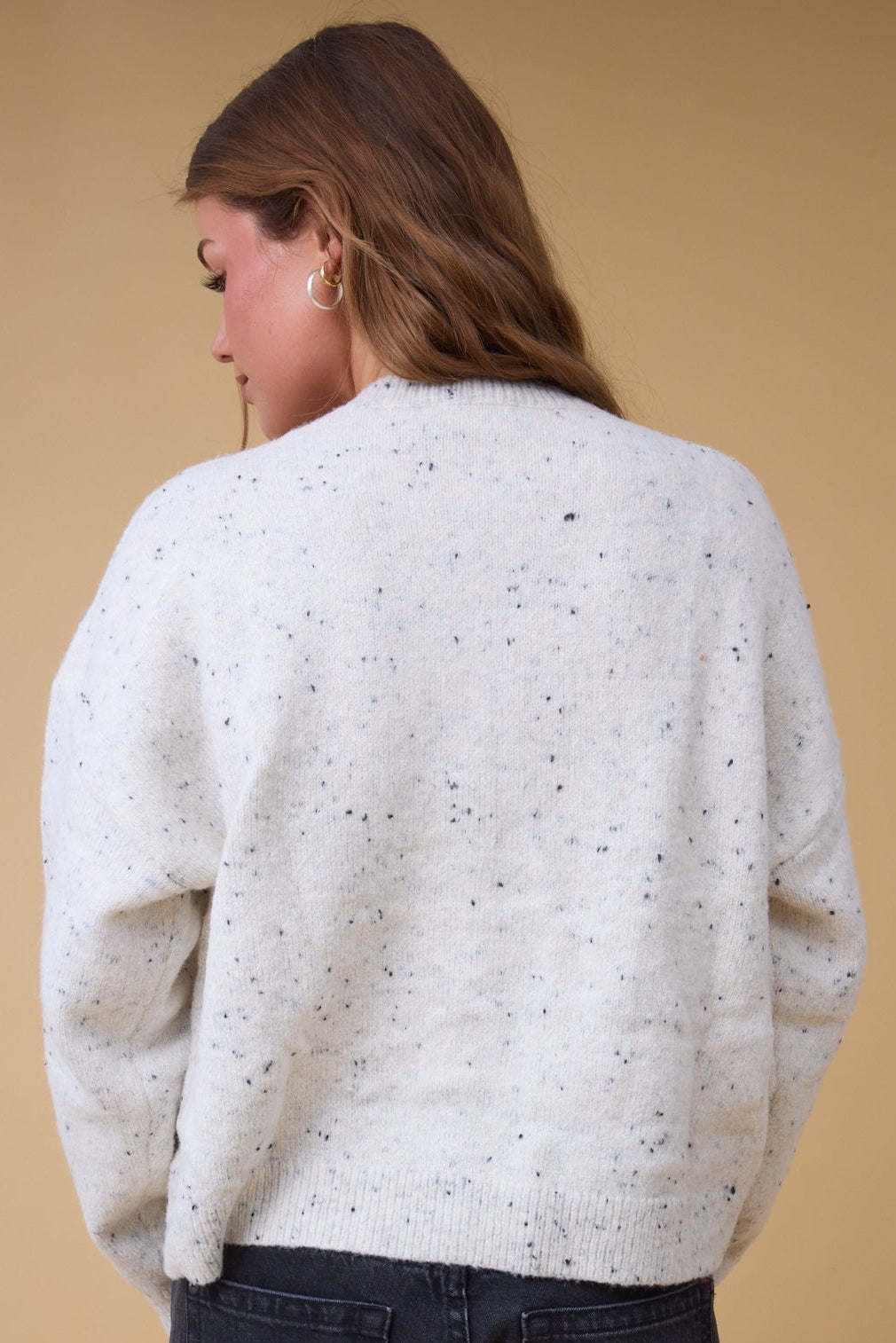 Back view of a woman wearing a cream flecked, loose fitting cardigan with long sleeves.
