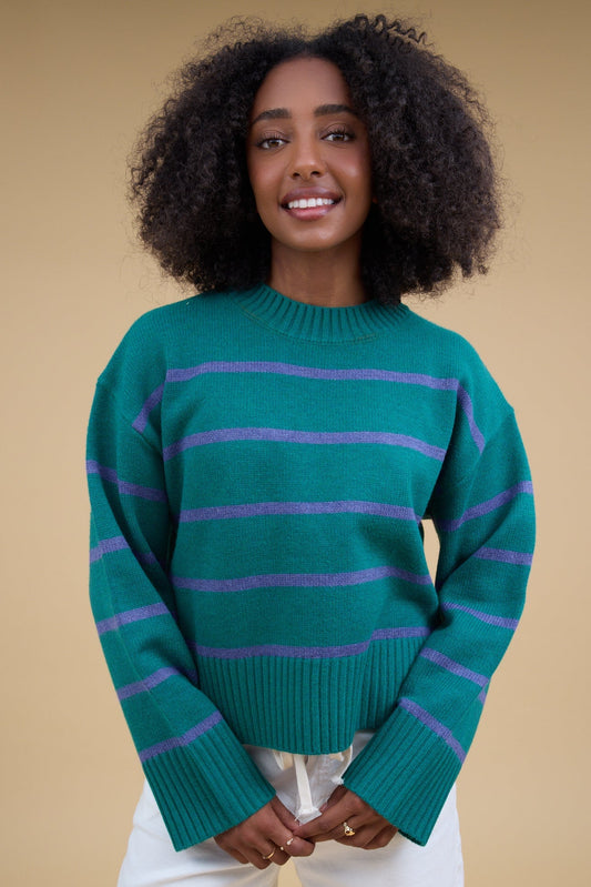 Young woman wearing a modest green & blue stripe pullover sweater with a wide ribbed crew neckline and long sleeves with wide loose ribb knit cuffs.