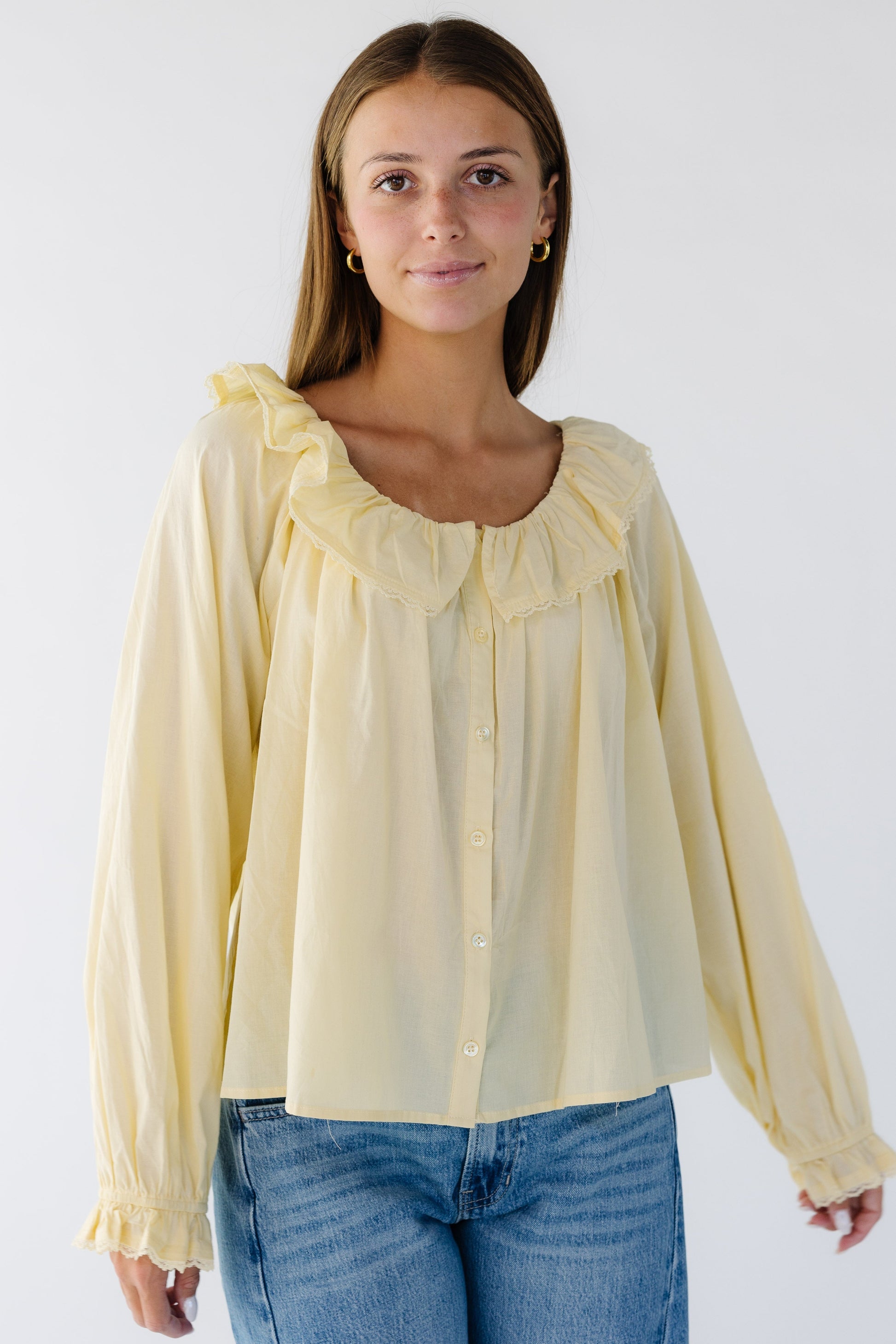 Young woman wearing a yellow peasant inspired loose fitting button down blouse with a scooped ruffled collar and long sleeves with ruffle cuffs.