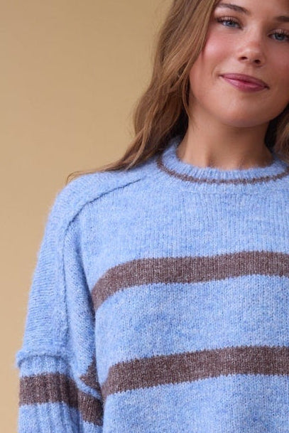 Close view of a young woman wearing a loose fitting stripe sweater in blue & brown stripe with exposed seams, drop shoulders and ribbed sleeves.