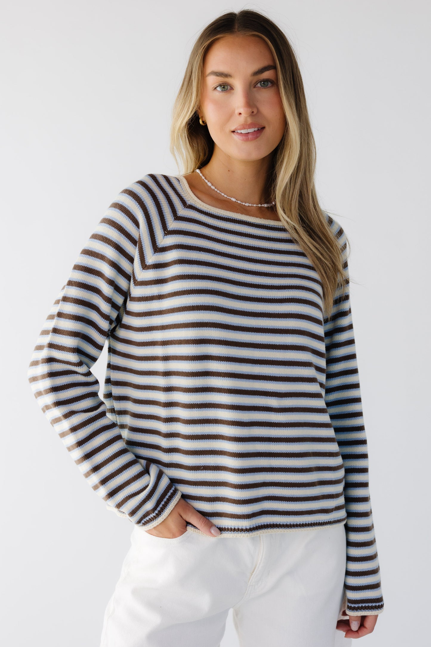 Young woman wearing a modest loose fitting sweater in cream, blue & brown stripe with long raglan sleeves and a wide neckline.