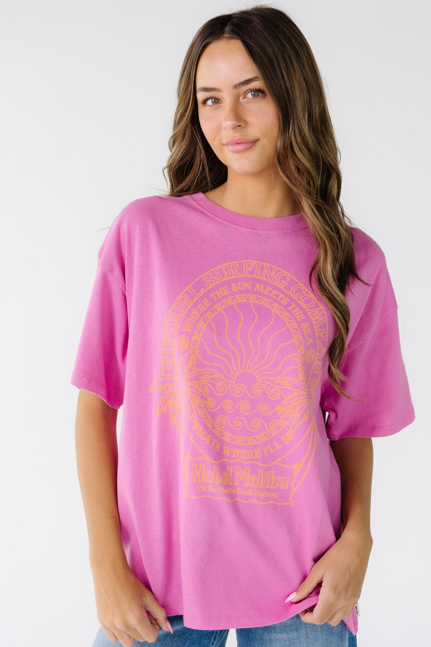 Woman wearing a modest bright pink t-shirt made by Rip Curl with short sleeves, crew neckline and a large front graphic in orange. 0MSWTE.