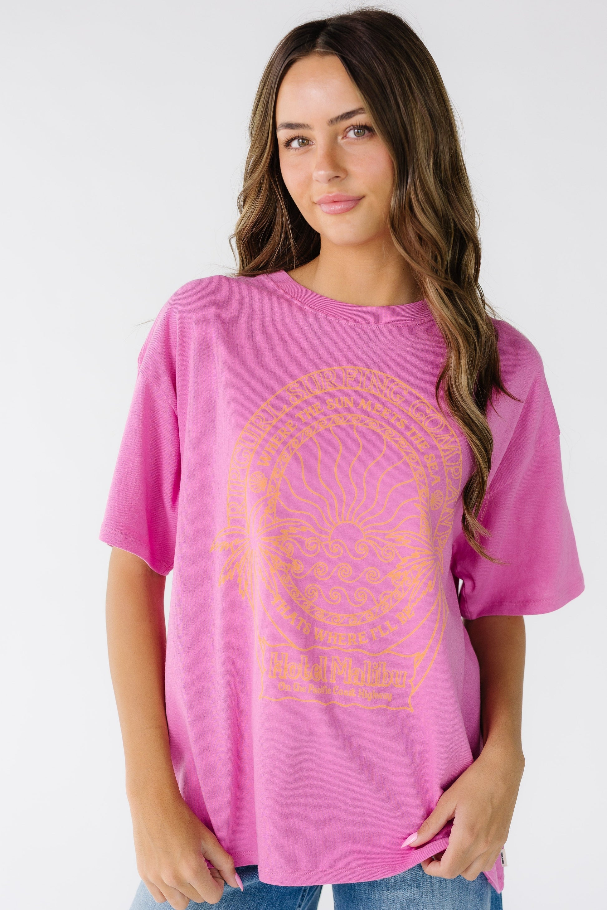 Woman wearing a modest bright pink t-shirt made by Rip Curl with short sleeves, crew neckline and a large front graphic in orange. 0MSWTE.