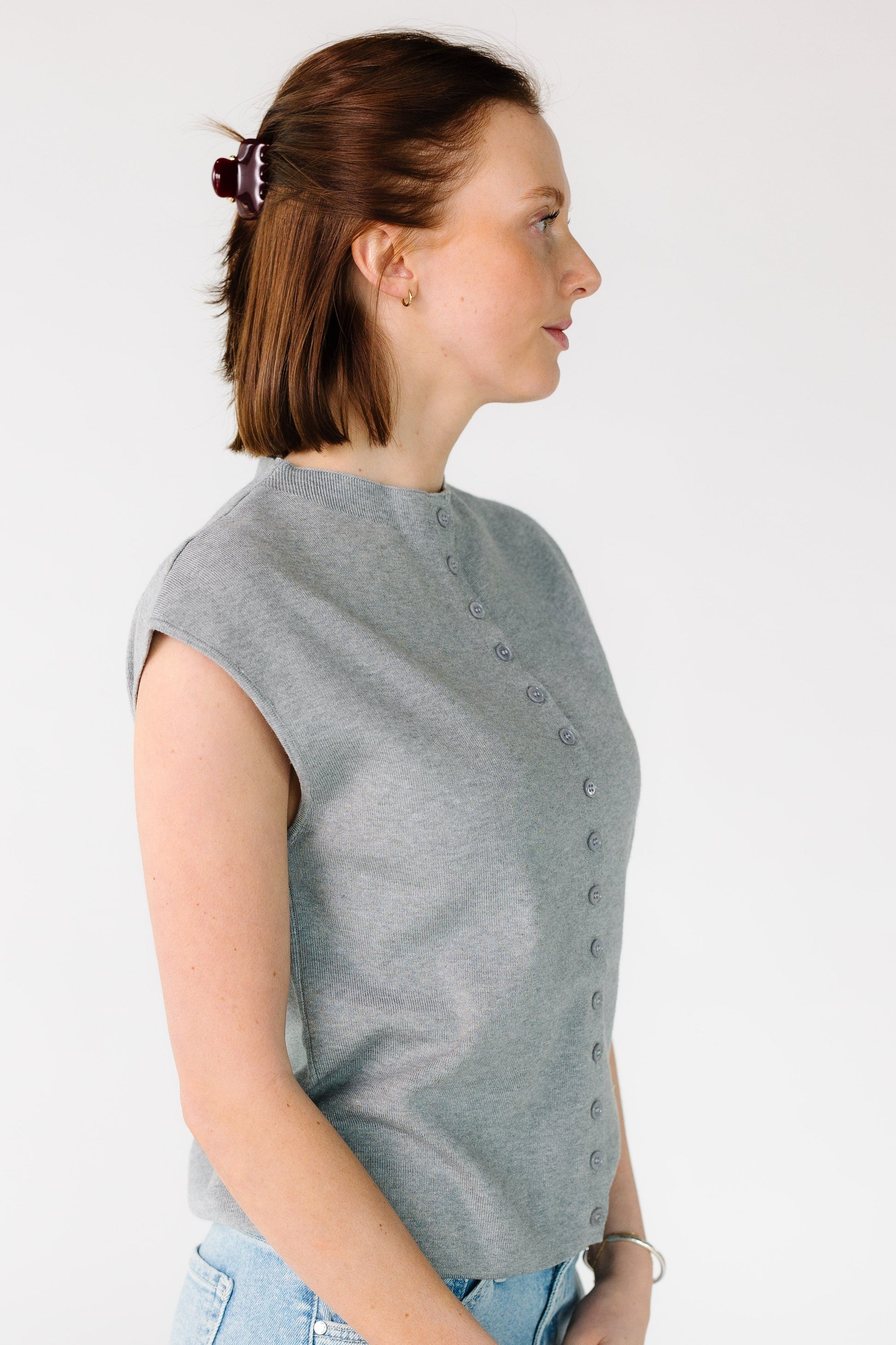 Side view of a woman wearing a sleeveless button down sweater vest in heather grey.