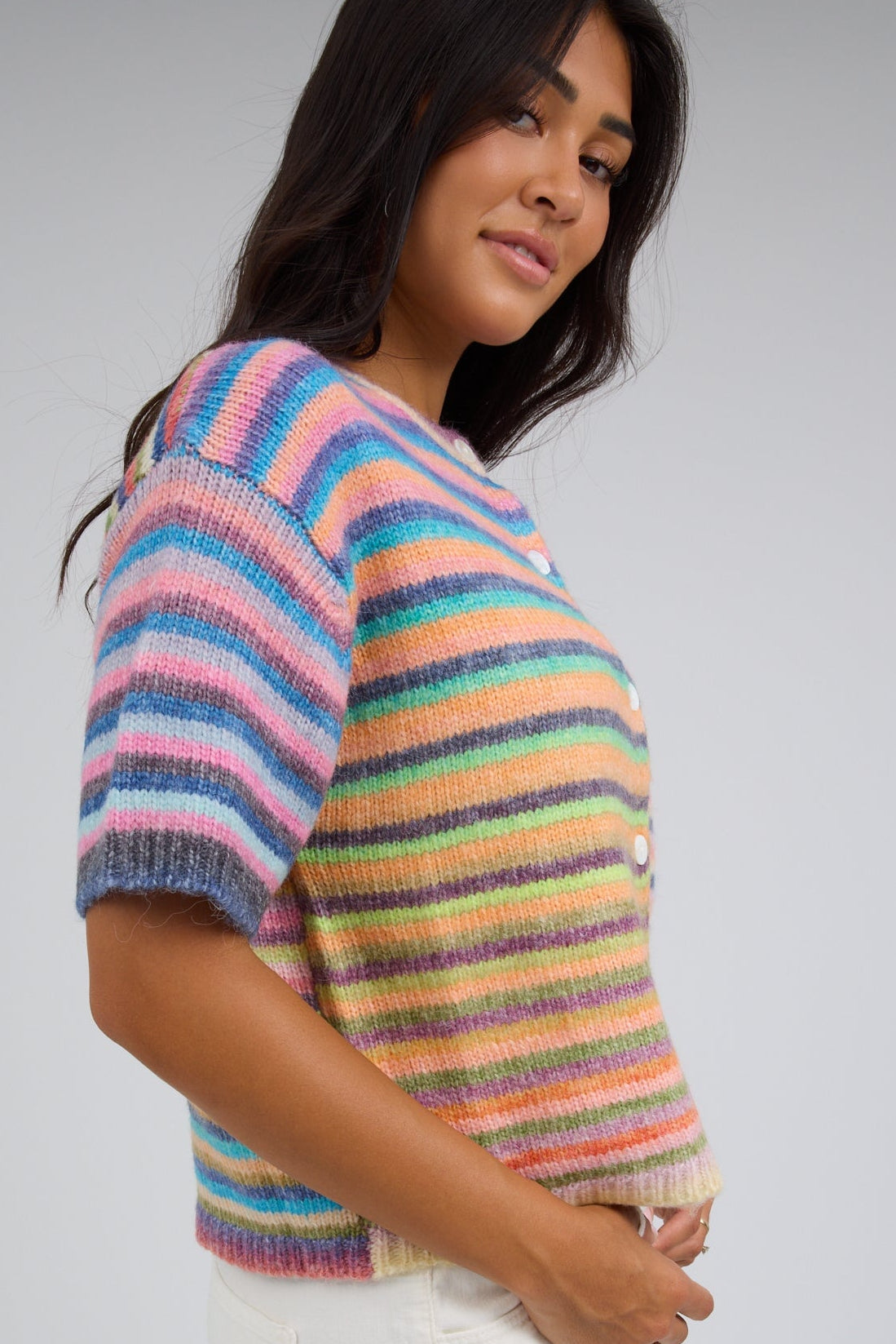 Side view of a young woman wearing a bright, rainbow striped cardigan with short sleeves.