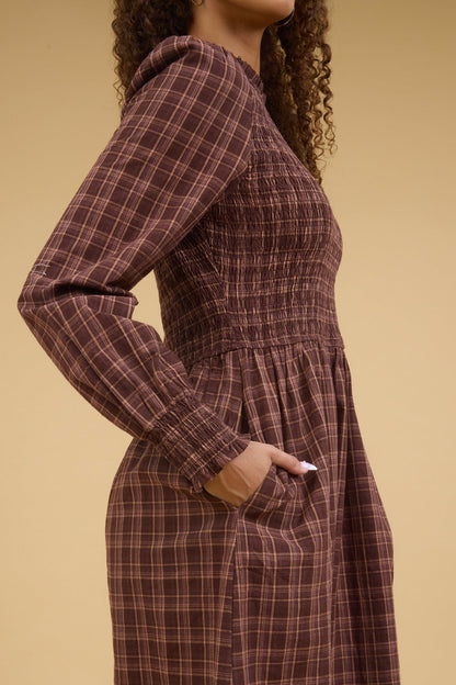 Close side view of a teenage girl wearing a brown plaid dress with long sleeves, smocked bodice and side pockets.