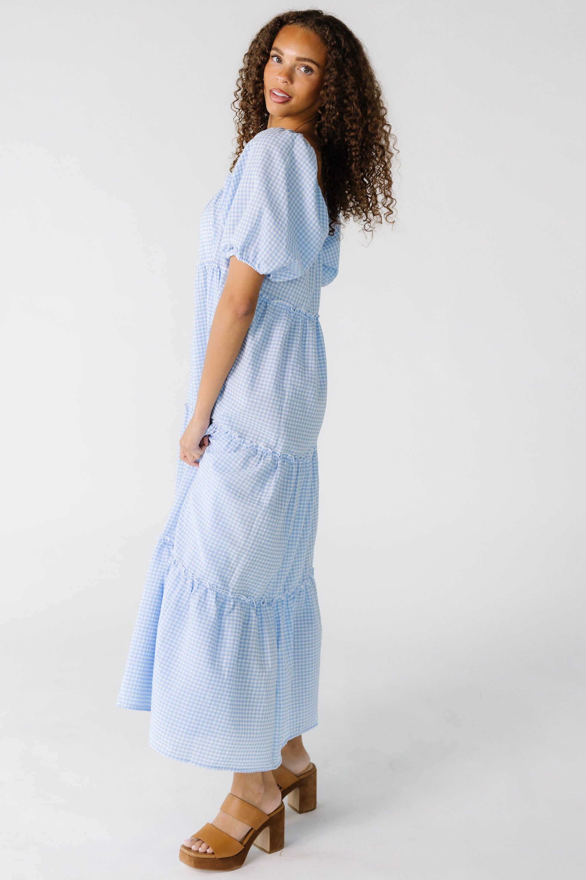 Side view of a teen girl wearing a light blue gingham tiered midi dress with large sleeves.