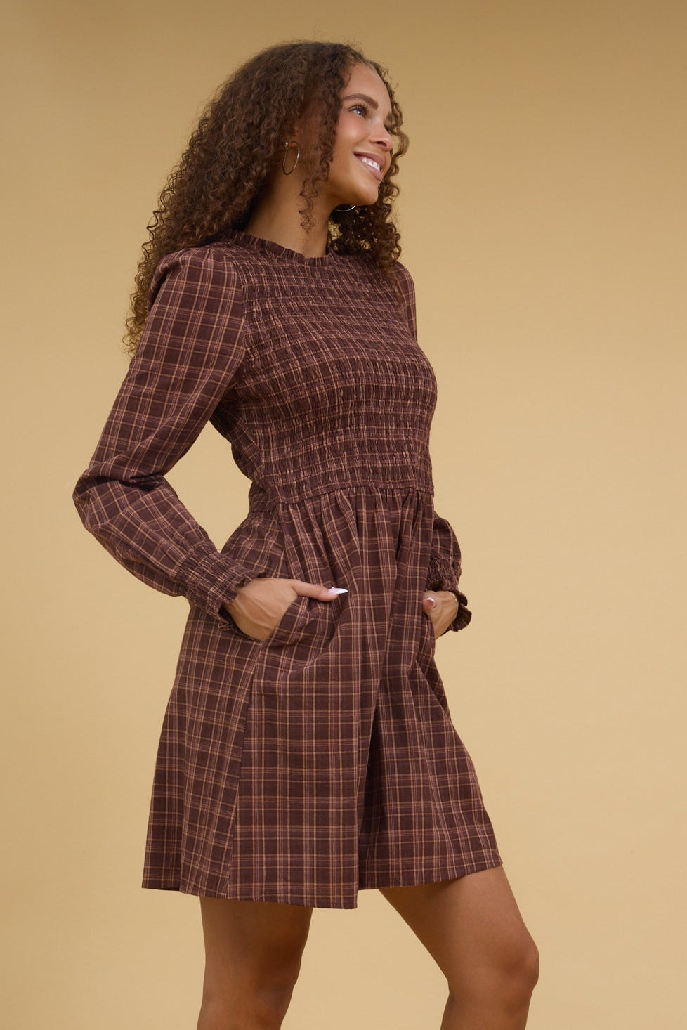 Teen girl wearing a brown plaid, short dress with side pockets, long sleeves and a smocked bodice.