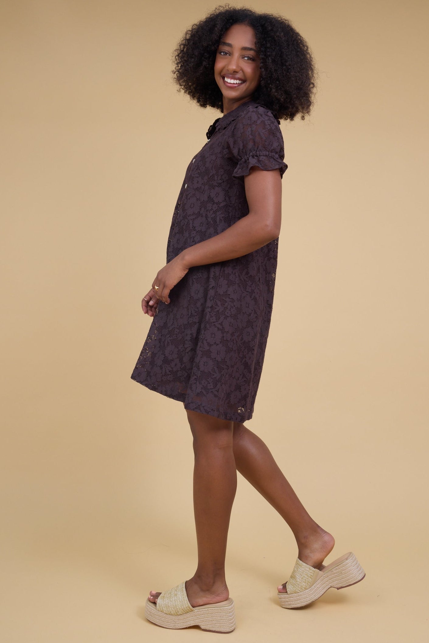 Side view of a woman wearing a brown all lace short dress with ruffle cuffed puff sleeves.