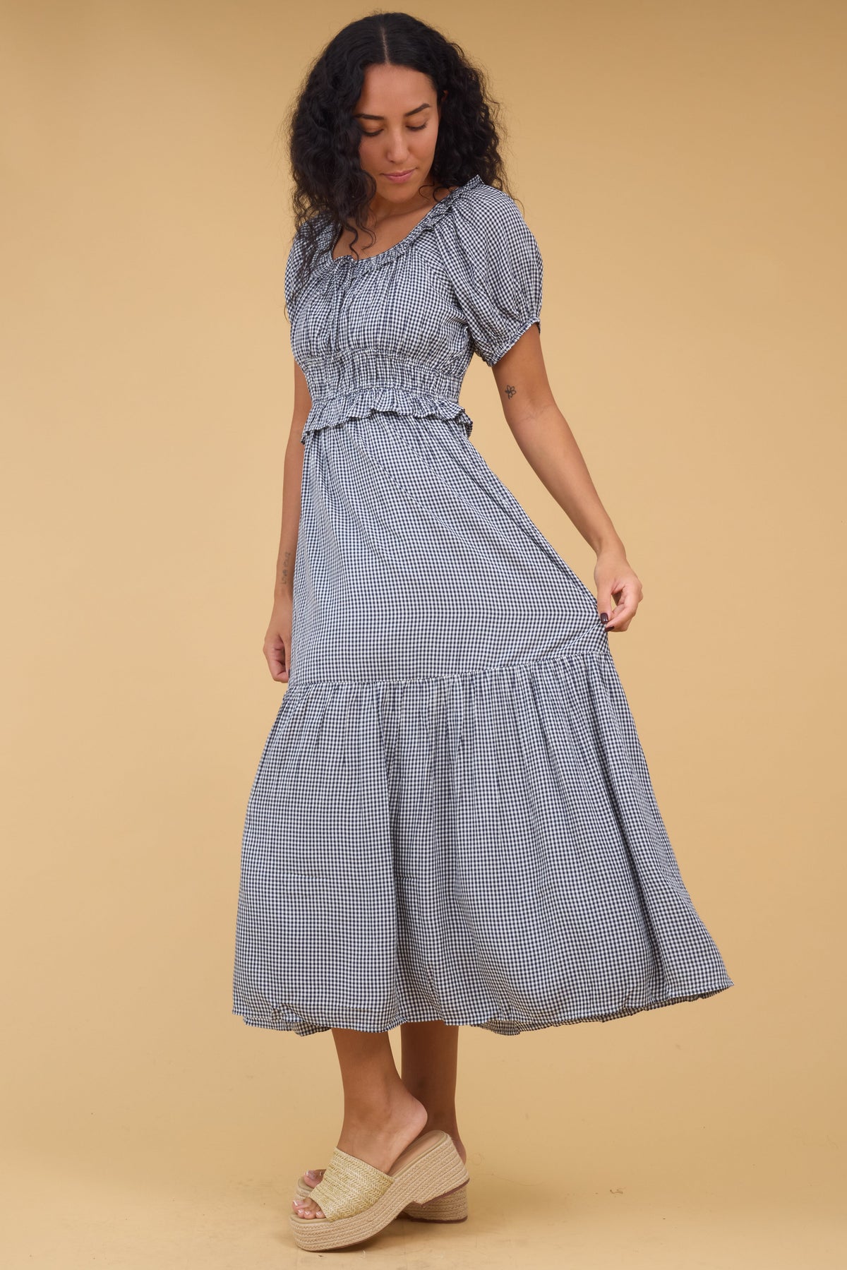 Young woman wearing a navy & white gingham midi dress with a wide bottom rufle, short puff sleeves, and a wide ruffled neckline.