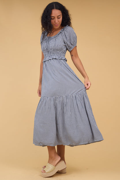 Young woman wearing a navy & white gingham midi dress with a wide bottom rufle, short puff sleeves, and a wide ruffled neckline.