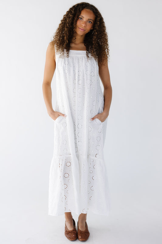 Young woman wearing a sleeveless,  white all eyelet midi-maxi dress with square neckline, side pockets and a wide bottom ruffle.