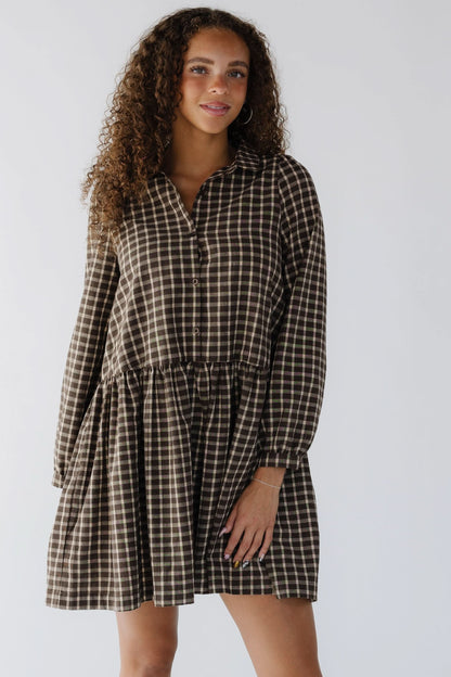 Young woman wearing a brown plaid, short dress that buttons down to the skirt, a pointed collar and long sleeves.