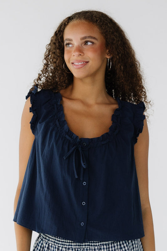 Young woman wearing a navy loose fitting, button down blouse with ruffled armholes and a scoop  & ruffled neckline with a tie.
