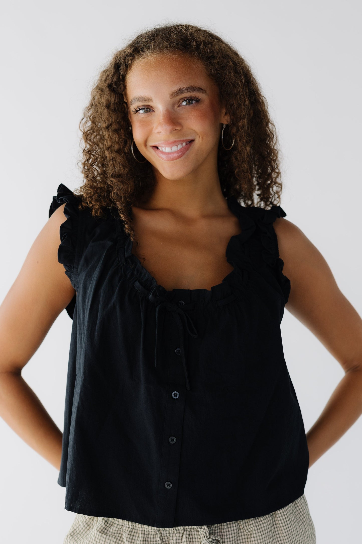 Young woman wearing a sleeveless loose fitting button down blouse with ruffled armholes and scooped ruffled neckline.