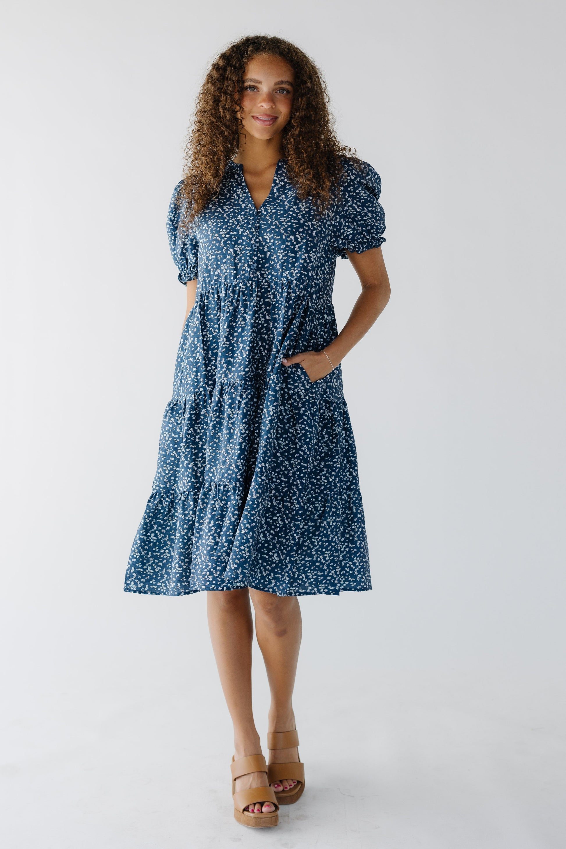 Teenage girl wearing a loose fitting tiered blue floral knee length dress with a V-neckline and short puff sleeves.