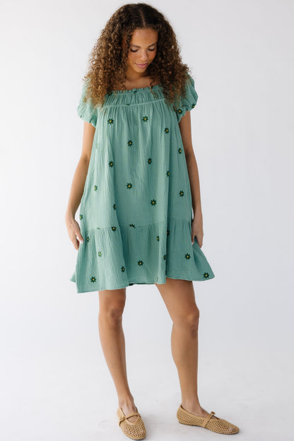 Teen girl wearing a short green loose fitting swing dress with embroidered green daisy's and short sleeves and a wide bottom ruffle.