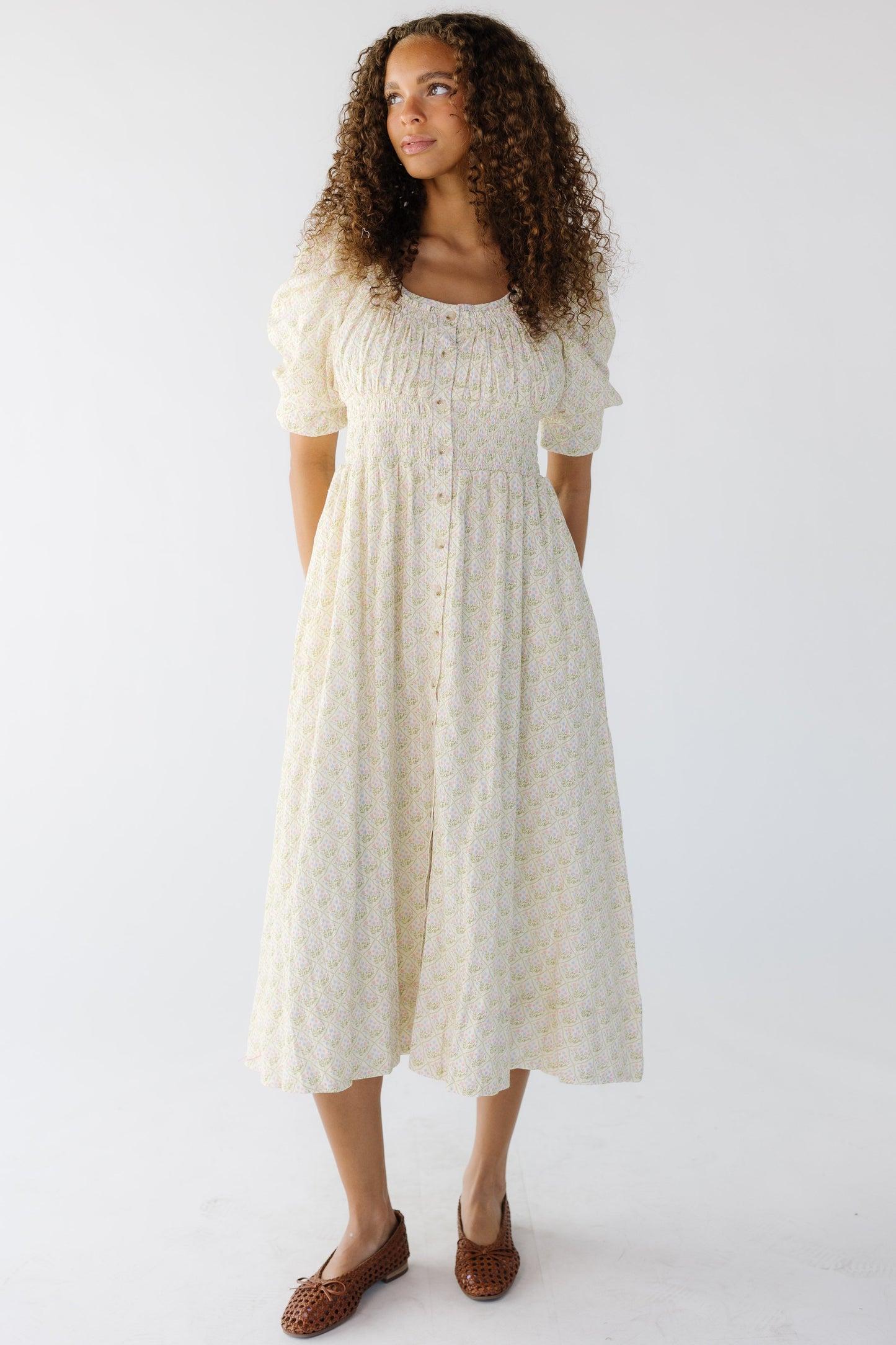 A young woman wearing a cream floral midi dress with short puff sleeves, a wide scoop neckline and a wide elastic wasitband.