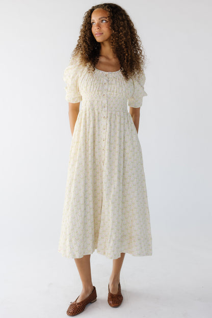 A young woman wearing a cream floral midi dress with short puff sleeves, a wide scoop neckline and a wide elastic wasitband.
