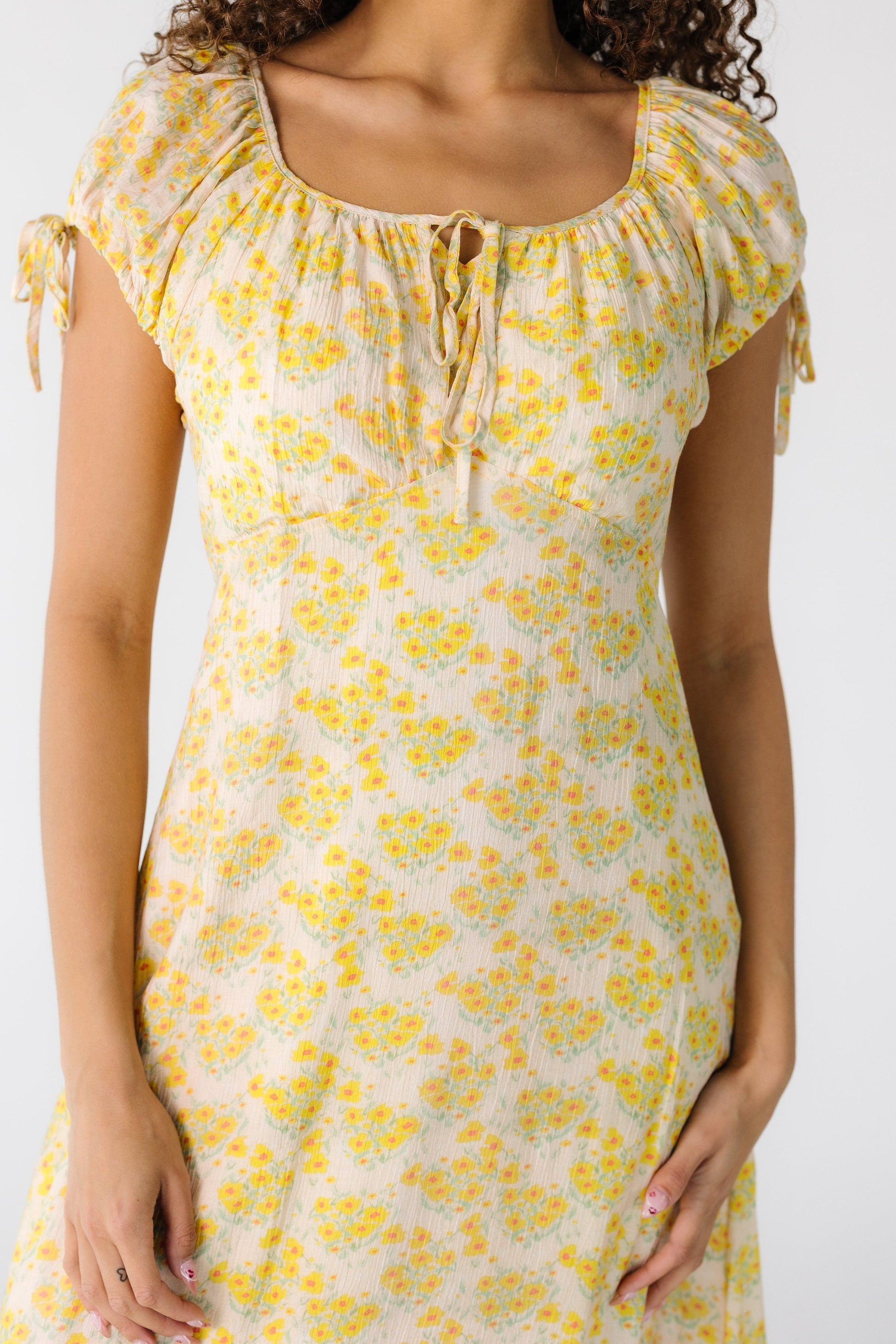 Close view of a teen girl wearing a yellow floral midi dress with a pointed empire waist square neckline with a tie and short puff sleeves with ties.