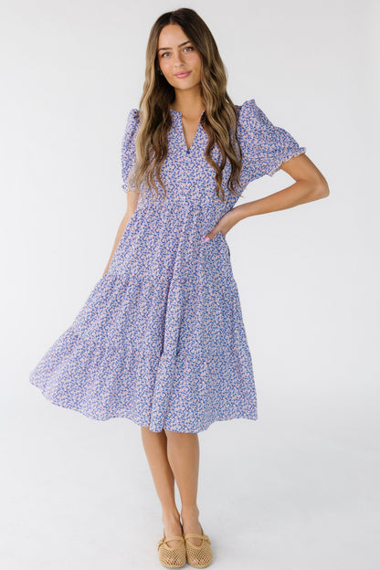 Young woman wearing a purple floral knee length tiered dress with a V-neckline and short puff sleeves.
