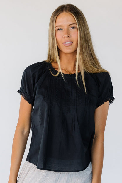 Young woman wearing a  black loose fitting blouse with a round lace trimmed neckline, short sleeves and embroidery, pleat and lace details.