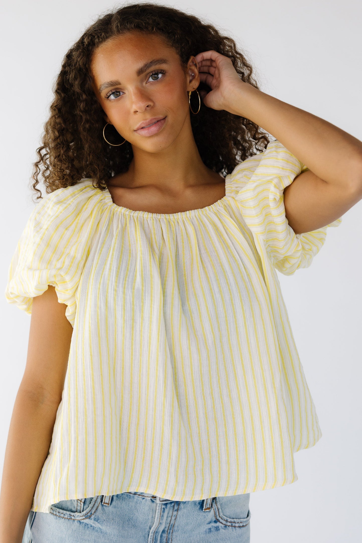 Young woman wearing a yellow and white vertical stripe loose fitting blouse with an elastic square neckline and large, puffy balloon sleeves.