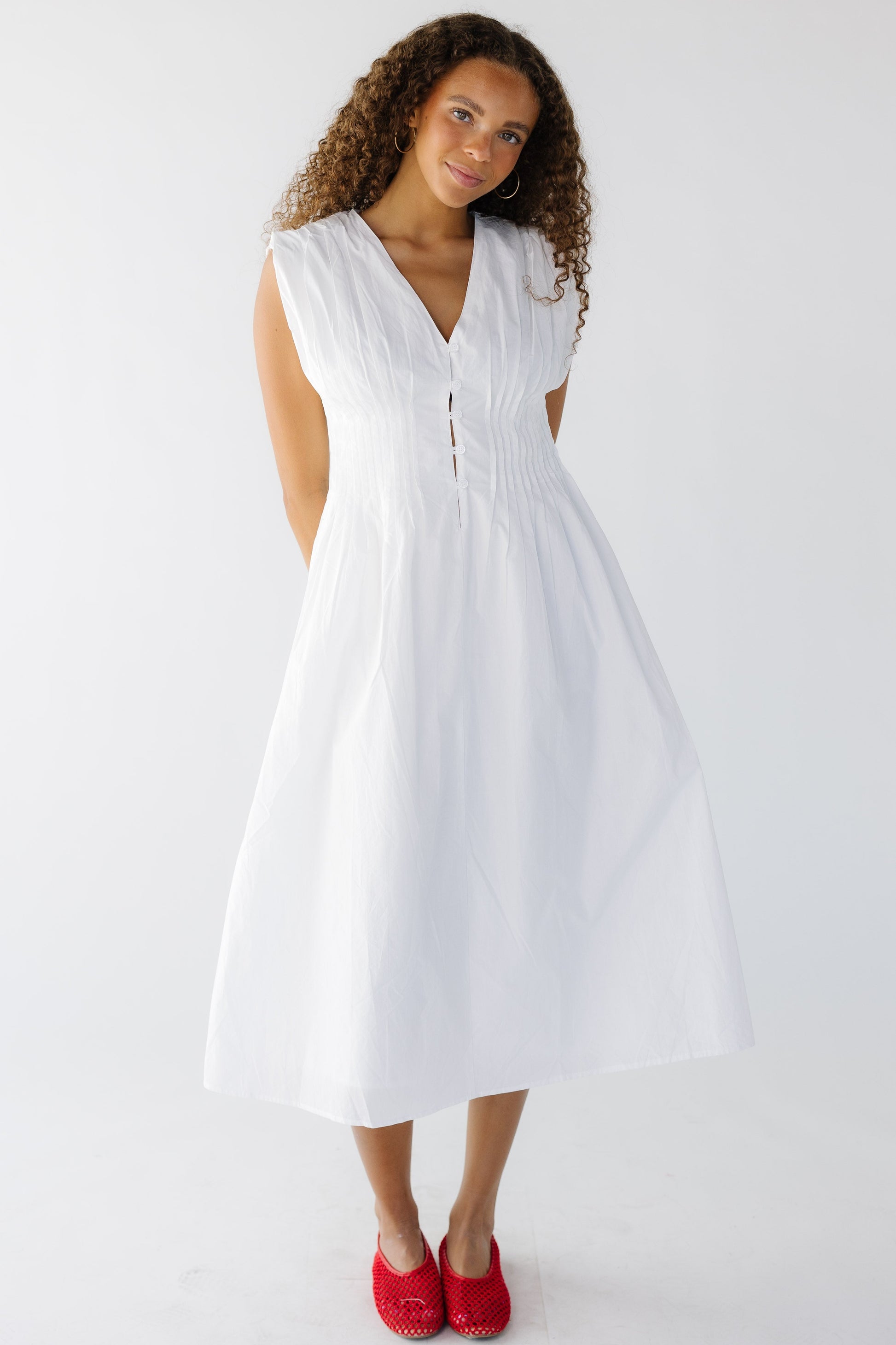 Teen age girl wearing a modest, sleeveless white midi dress with a V-neckline and pleats all around the waist a full A-line skirt.