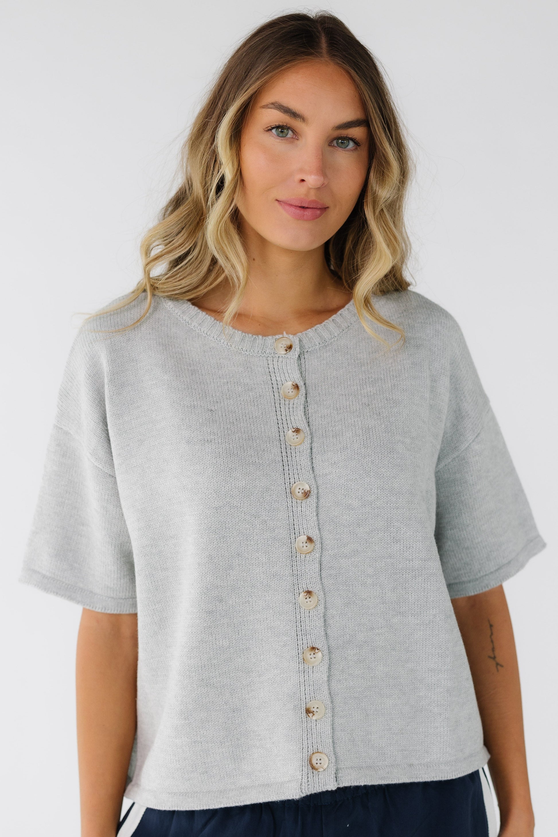 Young woman wearing a grey button down cardigan with a crew neckline, short sleeves and a boxy cut.  
