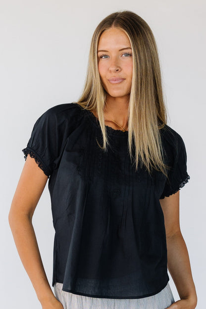 Young woman wearing a black flowy top with short lace hemmed sleeves and a round lace trimmed neckline with embroidery, lace and pleat details.