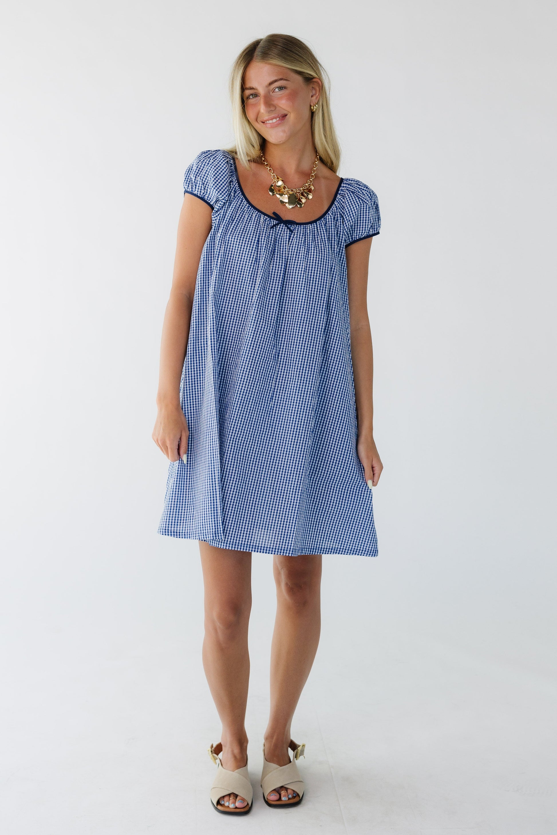 Young woman wearing a blue gingham dress cut above the knee with short puff sleeves and a wide scoop neckline.