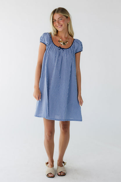 Young woman wearing a blue gingham dress cut above the knee with short puff sleeves and a wide scoop neckline.